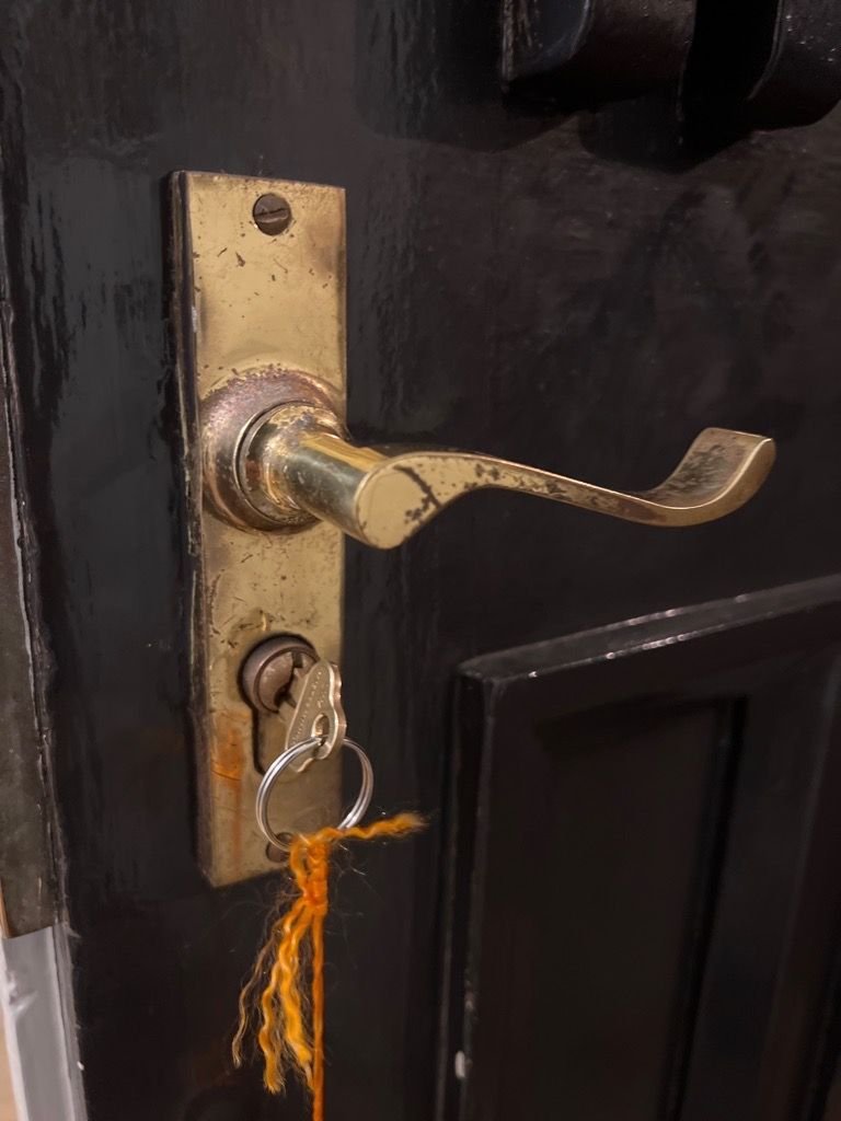 Old mortice lock on a black door with a key stuck in the barrel and an orange string attached to the keyring.