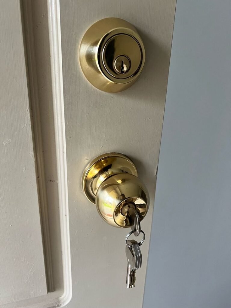 Close-up of polished brass deadbolt and doorknob lock on a white door with keys inserted.