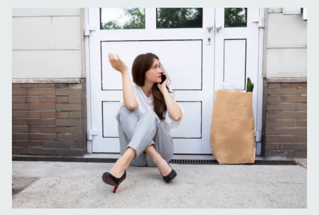 Woman sitting outside a closed door, talking on the phone with a grocery bag beside her.