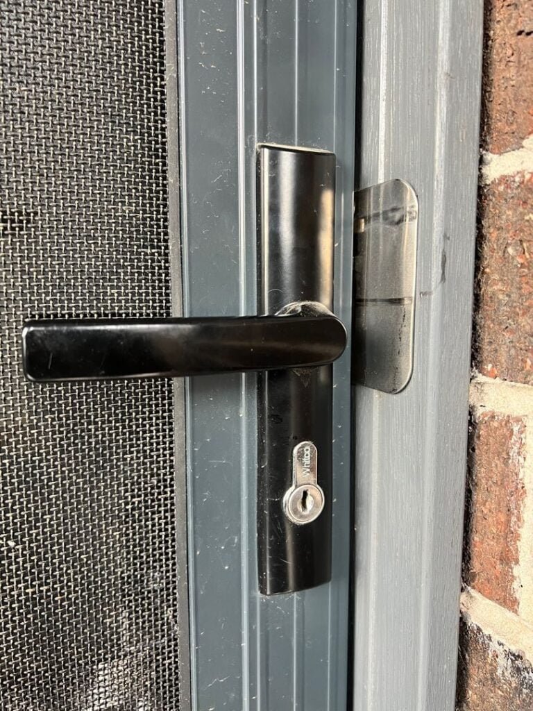 Close-up of a screen door lock and handle by Whitco on a metal-framed door next to a brick wall.