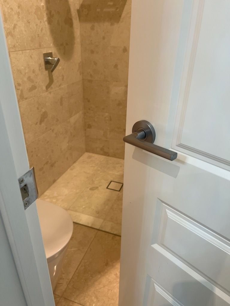 A partially open white door showing a bathroom with beige marble tiles and a silver showerhead.